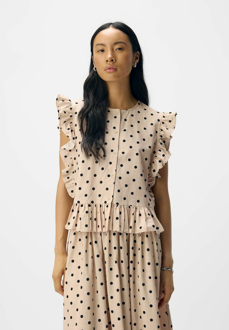 Robe rose clair à pois noirs, avec des manches volantées et une taille peplum. Texture lisse et boutons visibles.