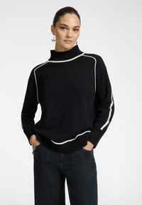 Maglione nero lavorato a maglia con collo alto, caratterizzato da strisce bianche lungo le cuciture. Indossato con jeans scuri, mani nelle tasche.