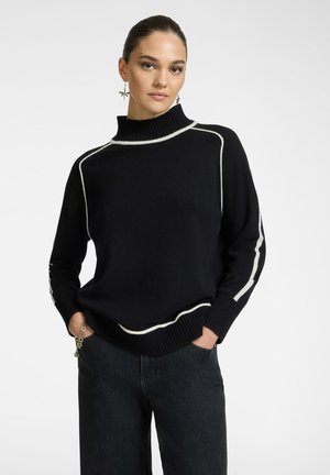Maglione nero lavorato a maglia con collo alto, caratterizzato da strisce bianche lungo le cuciture. Indossato con jeans scuri, mani nelle tasche.