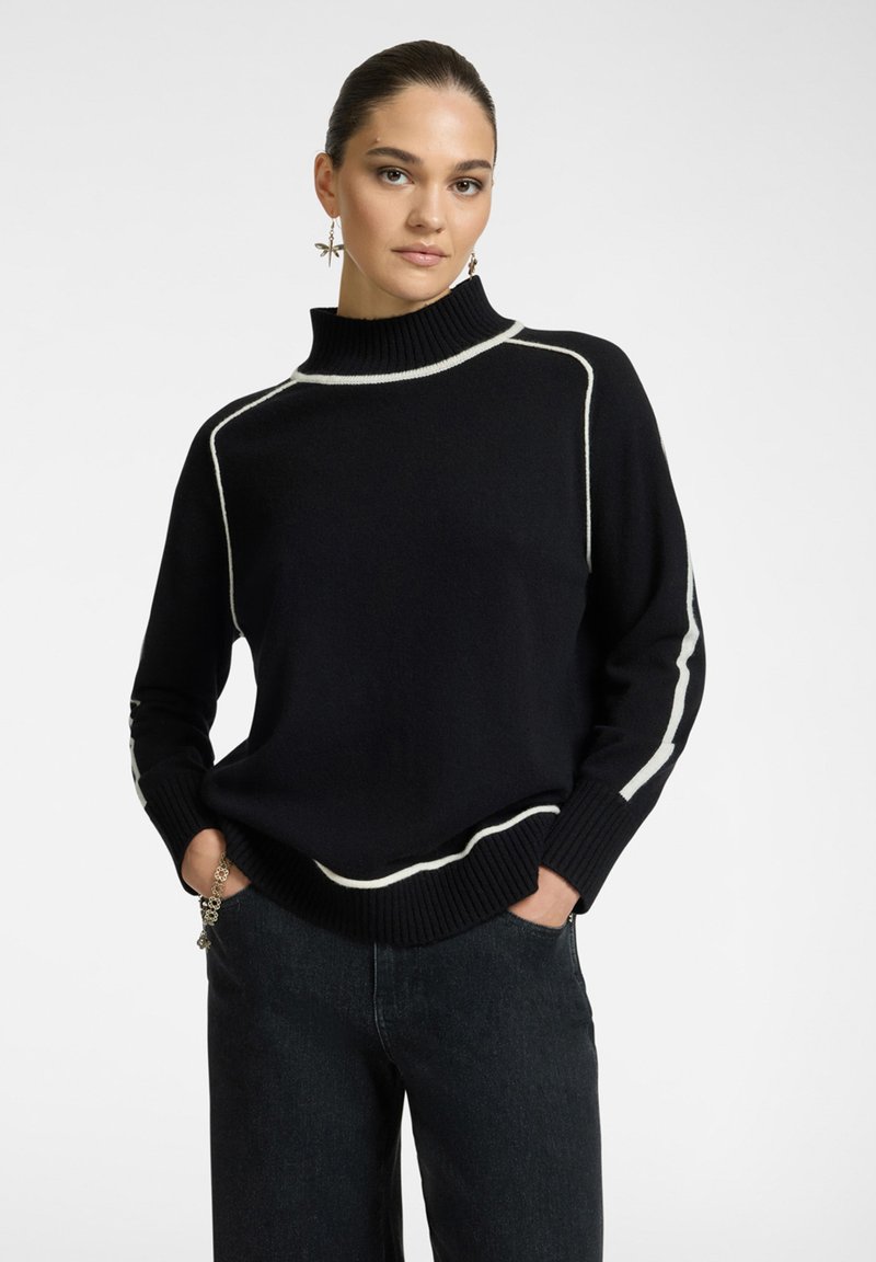 Maglione nero lavorato a maglia con collo alto, caratterizzato da strisce bianche lungo le cuciture. Indossato con jeans scuri, mani nelle tasche.