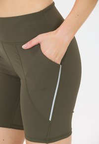 Läheltä kuvattuna henkilön käsi on oliivinvihreiden urheilushortsien taskussa, joissa on heijastava raita sivulla.