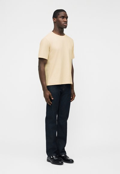 Νέος άνδρας που στέκεται, φορόντας μπεζ κοντομάνικο μπλουζάκι, σκούρα μπλε τζιν και μαύρα δερμάτινα παπούτσια μπροστά σε απλό λευκό φόντο.