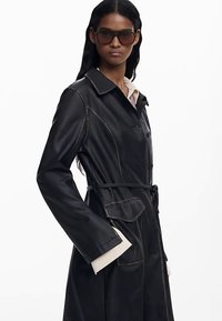 Černý kožený kabát s límcem, zapínáním na knoflíky a opaskem. Má kontrastní prošívání a detaily kapes. Hladká textura.