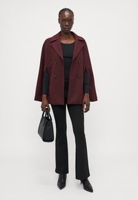 Burgundy dubbelknäppt cape-kappa med breda slag, kombinerad med en svart topp, utsvängda byxor och en liten svart handväska. Slät tygkvalitet.