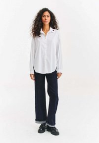 Bílá košile na knoflíky s dlouhými rukávy, kombinovaná s tmavými širokými džínami vyhrnutými na lemech a černými platformovými botami.