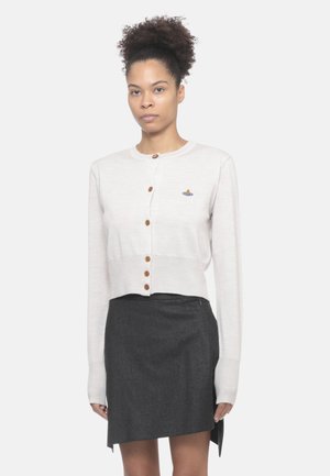 Cardigan corto grigio chiaro con colletto rotondo, bottoni marroni e un piccolo motivo ricamato blu e oro. Indossato sopra una gonna grigio scuro.