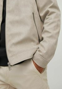 Beige suede jacket with zip fastening and ribbed hem. Features a side pocket and smooth texture, showcased partially with hands in pockets.