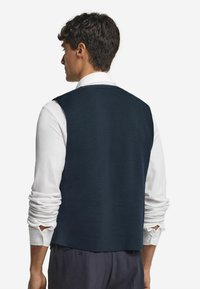 Gilet blu realizzato in tessuto texture con taglio dritto, indossato sopra una camicia bianca a maniche lunghe. La vista posteriore non mostra hardware o dettagli visibili.