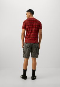Rød kortærmet T-shirt med sort geometrisk mønster, kombineret med grå cargo shorts. Modellen står med ryggen til kameraet iført sorte sneakers.