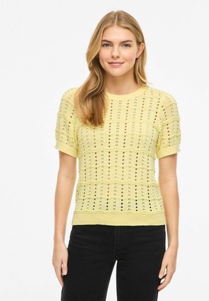 Jeune femme aux longs cheveux blonds portant un haut léger en maille jaune pâle à manches courtes avec des motifs ajourés et un pantalon noir, debout devant un fond blanc.