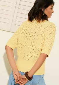 Suéter amarillo de punto con mangas cortas, presenta patrones de diamante, tejido texturizado y dobladillo acanalado. Combinado con jeans y un brazalete marrón.
