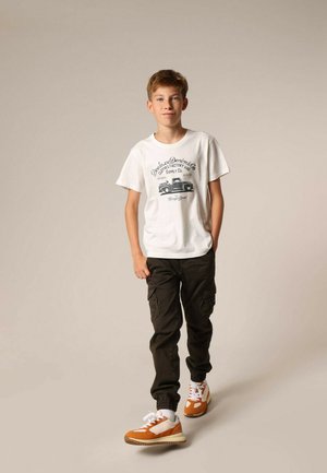 Witte T-shirt met zwart grafisch ontwerp, olijfgroene cargo-broek en oranje-witte sportschoenen. Kind staat tegen een neutrale achtergrond.