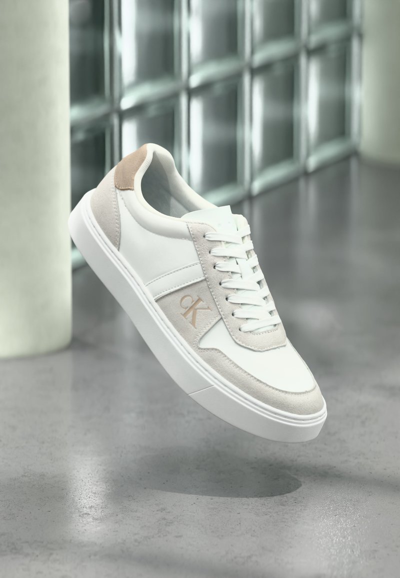Λευκό και μπεζ sneaker χαμηλού τύπου με κορδόνια και λογότυπο CK, αιωρούμενο πάνω από γυαλισμένο τσιμεντένιο πάτωμα με φόντο τοίχο από γυάλινα τούβλα.