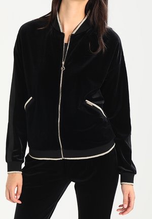 Kvinde iført en sort velourjakke med lynlås, med lyse kanter på krave, manchetter, kant og lynlåslommer, kombineret med matchende bukser.