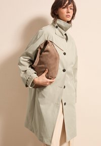Lichtgroene trenchcoat met zwarte knopen, brede kraag en ribgebreide manchetten, gecombineerd met een zachte bruine suède schoudertas.