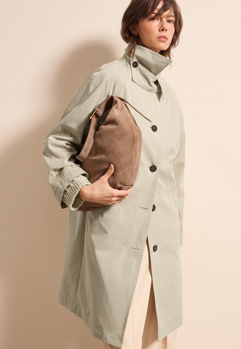 Lichtgroene trenchcoat met zwarte knopen, brede kraag en ribgebreide manchetten, gecombineerd met een zachte bruine suède schoudertas.