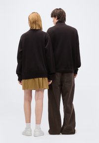 Δύο άτομα στέκονται με την πλάτη, και οι δύο φορούν μαύρα φούτερ· το ένα φορά κίτρινη φούστα καρό με λευκά αθλητικά παπούτσια, το άλλο καφέ παντελόνια.