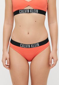 Calvin Klein juostu rinkinys, kuriame yra koralų spalvos trumpa palaidinė ir derančios kelnaitės. Juosmuo juodas su kontrastingais baltais prekės ženklo užrašais.