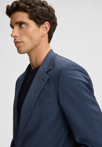 Marineblazer mit strukturiertem Gewebe, strukturiertem Revers und einer einzelnen Brusttasche, getragen über einem dunklen Hemd. Professionelles, tailliertes Design.