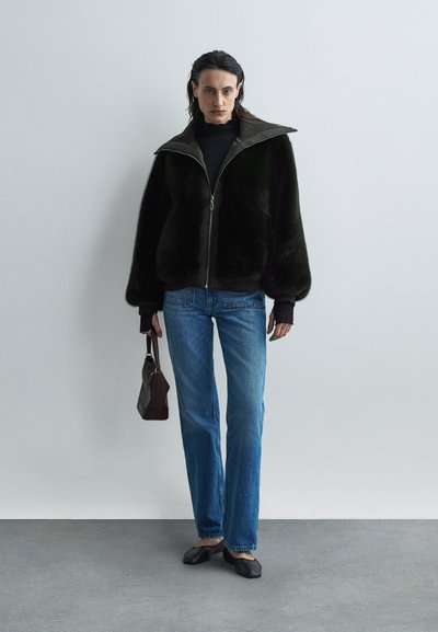 Giacca in pelliccia sintetica verde scuro con ampio colletto, chiusura con zip, abbinata a jeans blu a gamba dritta e ballerine nere, mentre tiene in mano una piccola borsa marrone.