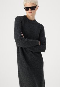 Καστόν φόρεμα σε μαύρο χρώμα με μακριά μανίκια, στρογγυλή λαιμόκοψη και ραβδωτή υφή. Το μοντέλο φοράει γωνιακά μαύρα γυαλιά ηλίου και στέκεται μπροστά σε ανοιχτό φόντο.