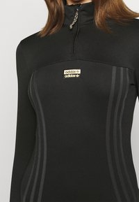 Černý sportovní top na zip s zlatým logem Adidas, texturovaná látka a tři tmavé pruhy po stranách pro detail.