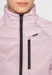 Giacca rosa chiaro realizzata in materiale lucido, con colletto alto, chiusura centrale a zip e tasca frontale con zip sul petto, con accenti neri.