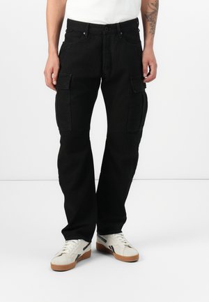 Schwarze Cargo-Hosen mit lockerer Passform, ausgestattet mit mehreren Taschen, Gürtelschlaufen und robustem Stoff. Kombiniert mit weißen Sneakers und einem schlichten Shirt.