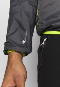 Veste imperméable grise avec des accents vert lime, arborant un logo sur la manche, et une poche zippée sur le pantalon noir.