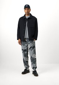 Fekete gombos gyapjú keverék dzseki mellzsebbel, szürke póló, szürke álcázott flísznadrág és fekete cipő szürke részletekkel.