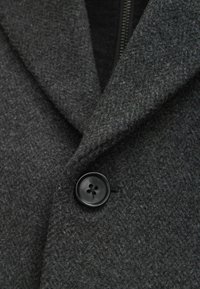 Abrigo de lana gris oscuro con una superficie texturizada, que cuenta con un botón negro de cuatro orificios y un cuello de chal. Primer plano de la tela y el herraje.