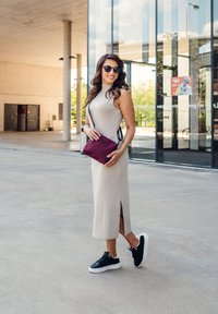 Beige bezrukávové šaty s bočním rozparkem, doplněné černými teniskami. Drží malou bordó crossbody tašku s ramenním popruhem.