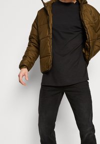 Chaqueta acolchada verde oliva con cremallera y capucha, con un diseño acolchado. Llevada sobre una camiseta negra y combinada con vaqueros negros.