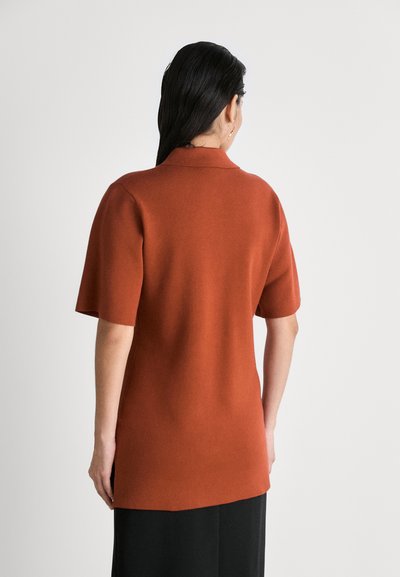 Haut à manches courtes, couleur rouille, avec un col, présentant une texture lisse et une coupe décontractée qui descend légèrement au-delà des hanches.