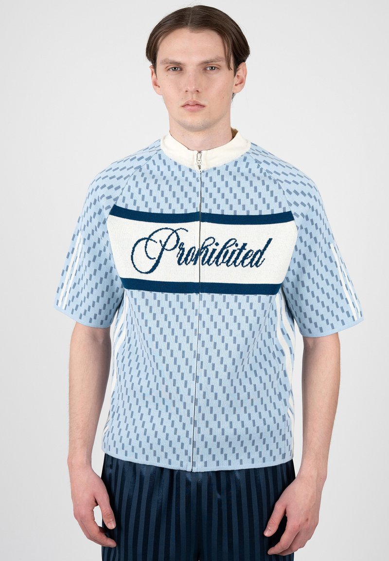 Jeune homme aux cheveux bruns et raides porte un haut bleu à manches courtes à motifs avec fermeture éclair, avec « Prohibited » écrit sur la poitrine, associé à un pantalon foncé rayé.