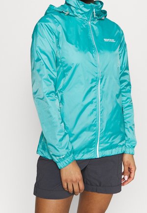 Veste légère zippée couleur sarcelle avec capuche portée avec un short sombre, montrée sur une personne du cou à mi-cuisse, debout immobile.