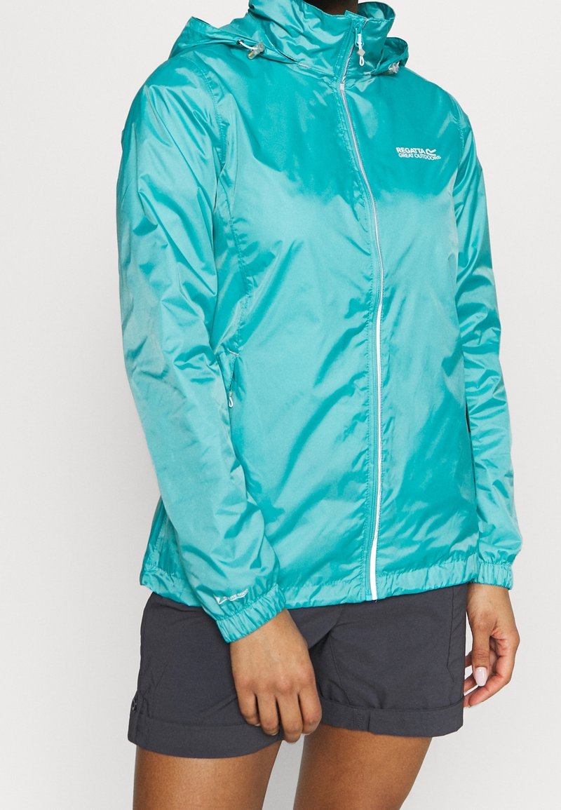 Veste légère zippée couleur sarcelle avec capuche portée avec un short sombre, montrée sur une personne du cou à mi-cuisse, debout immobile.