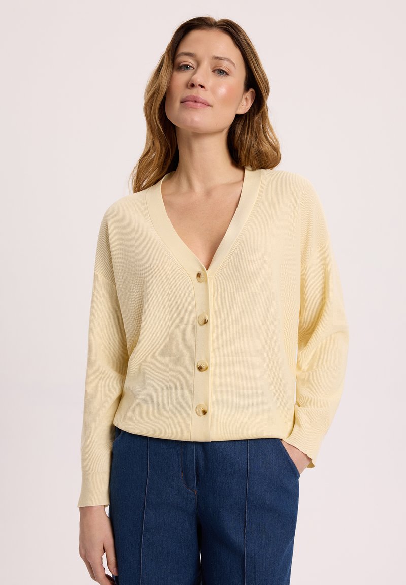 Kobieta o jasnobrązowych włosach nosi bladożółty sweter na guziki i niebieskie spodnie, stoi z rękami w kieszeniach na jednolitym tle.