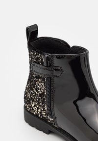 Botín de tobillo de charol negro con panel texturizado brillante en negro y dorado. Cuenta con una cremallera lateral y una pestaña de anilla para facilitar su uso.