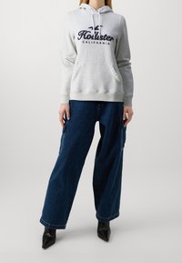 Felpa grigia in cotone con tasca a marsupio e stampa blu navy, abbinata a pantaloni denim blu scuro larghi e décolleté nere con punta a punta.