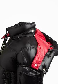 Giacca a vento nera lucida con colletto alto e rivestimento interno rosso, dotata di bottoni automatici, zip e una tracolla nera con l'etichetta "JACKIT."