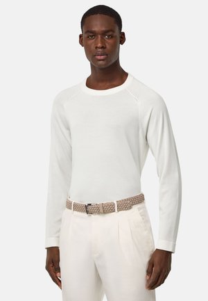 Camicia bianca a maniche lunghe con maniche raglan, abbinata a pantaloni crema e una cintura intrecciata marrone chiaro con fibbia metallica.