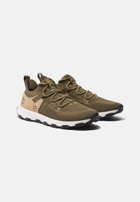 Oliivinvihreät neulottu sneakerit ruskeilla yksityiskohdilla, rakenneverkosta valmistettu yläosa, nauhoitusjärjestelmä ja valkoiset välipohjat, joissa on kestävä musta ulkopohja.