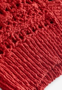 Tissu tricoté rouge avec un motif texturé, comportant des mailles ouvertes et un bord côtelé. Le matériau semble épais et confortable.