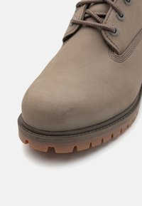 Bota de ante marrón con punta redonda, diseño con cordones, ojales grises y una suela de goma resistente con prominentes tacos para tracción.
