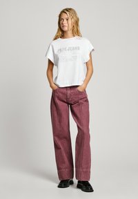 T-shirt blanc court avec impression de texte, associé à un jean large taille haute bordeaux. Modèle portant des mocassins noirs. Fond neutre.