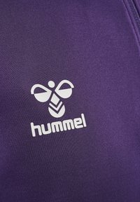 Fioletowy strój sportowy z dużym białym logo przedstawiającym stylizowaną pszczołę oraz napisem "hummel" poniżej. Gładka tekstura, minimalistyczny design.