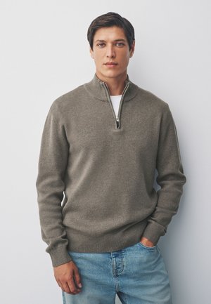 Giovane uomo con capelli castani che indossa un maglione grigio con zip sopra una camicia bianca e jeans azzurri, in piedi davanti a uno sfondo bianco.