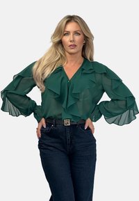 Green sheer blouse with ruffled accents and long, bell sleeves, paired with dark denim high-waisted jeans and a belt with a metallic buckle.