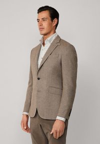 Brauner Tweed-Blazer mit Reverskragen, einreihiger Knopfschluss und zwei Fronttaschen, getragen über einem karierten Hemd.